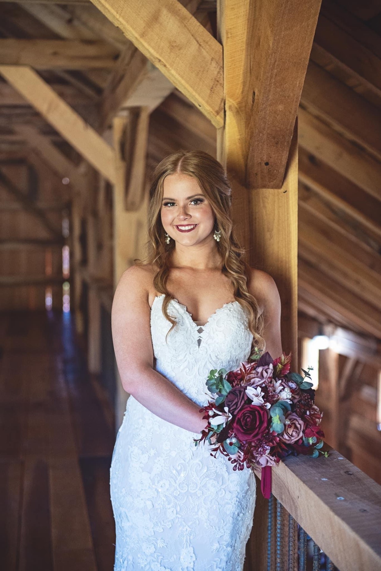 Bride in strapless lace gown holding burgundy bouquet in barn loft